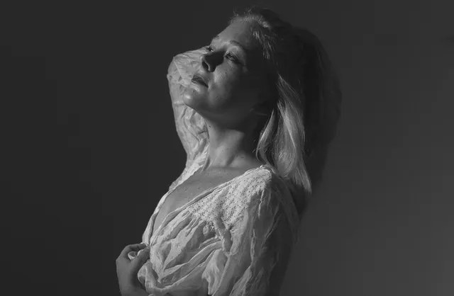 Blond model with sensual gaze in transparent white top kneeling during sexy photoshoot